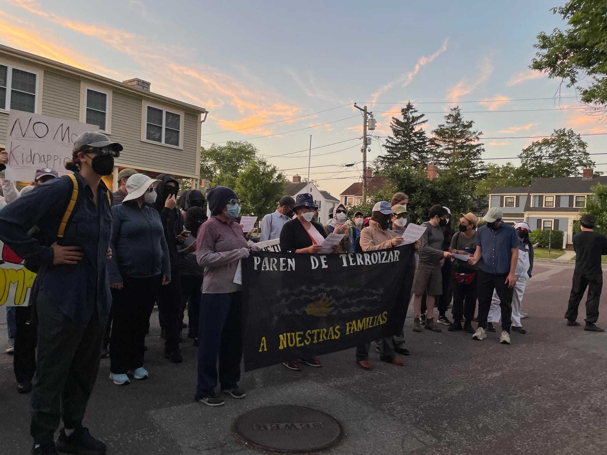 Protesters standing on the street in front of Governor Healey's home hold a sign reading "Stop terrorizing our families" in Spanish.