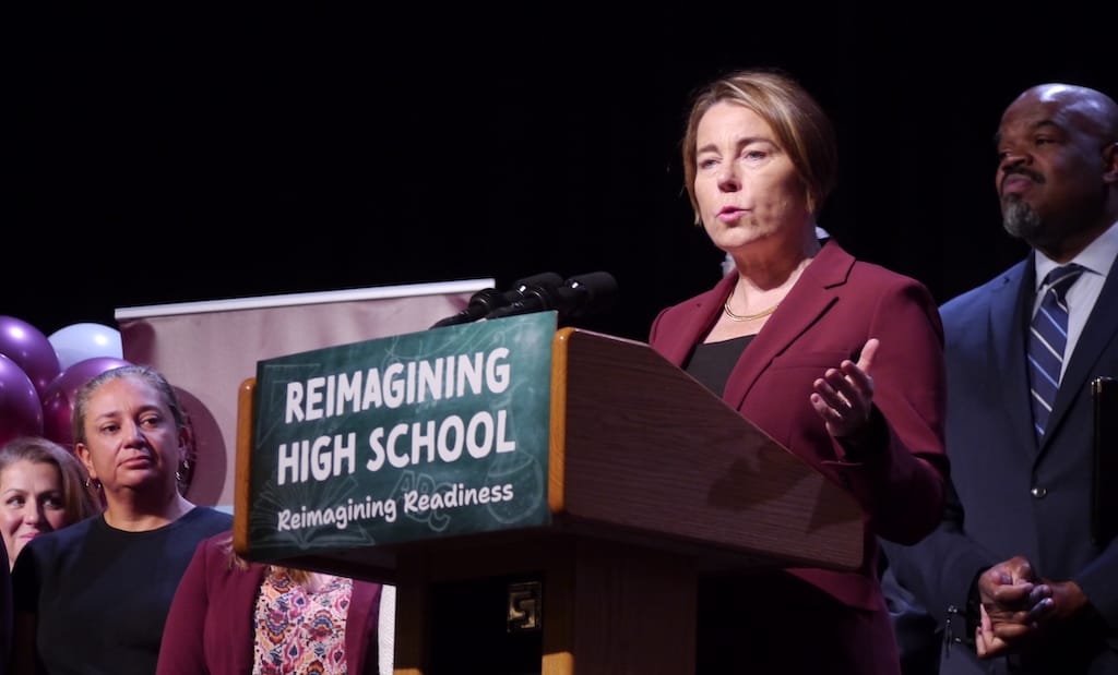 Governor Maura Healey speaks from a podium at Dedham High School as Education Secretary Paul Tutwiler and others look on.