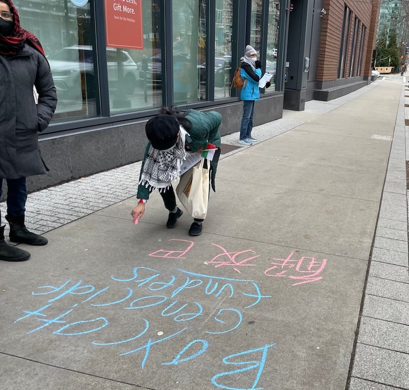 A demonstrator chalks "Boycott genocide funding" in Roman script and Chinese characters on the sidewalk outside Capital One's Seaport Boulevard location.