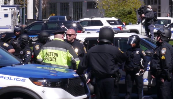 Boston police officers gather at the site of a Northeastern University student protest against Israel's siege of Gaza while a camera operator looms in background.