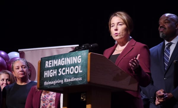 Governor Maura Healey speaks from a podium at Dedham High School as Education Secretary Paul Tutwiler and others look on.
